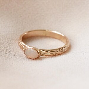 Pu&ograve; includere: Un delicato anello d'oro con una pietra di opale bianca incastonata al centro. La fascia &egrave; decorata con un motivo floreale ripetitivo.
