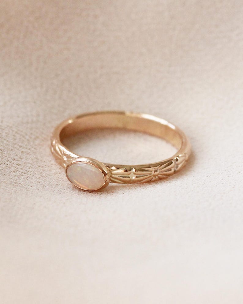 Pu&ograve; includere: Un delicato anello d'oro con una pietra di opale bianca incastonata al centro. La fascia &egrave; decorata con un motivo floreale ripetitivo.