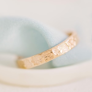 Peut inclure: Une bague anneau en or présentant un motif floral gravé répétitif sur toute la surface extérieure. Le métal possède une finition polie qui reflète la lumière, mettant en valeur les détails complexes des pétales sculptés sur la circonférence du bijou.