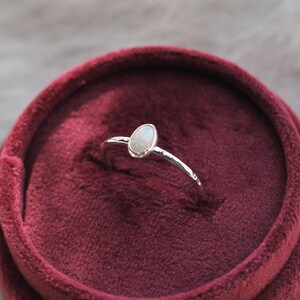 May include: A silver ring featuring a small, oval-shaped iridescent opal stone set in a smooth bezel. The ring band has a textured, hammered finish and is displayed upright in a deep red, circular velvet-lined jewelry box.
