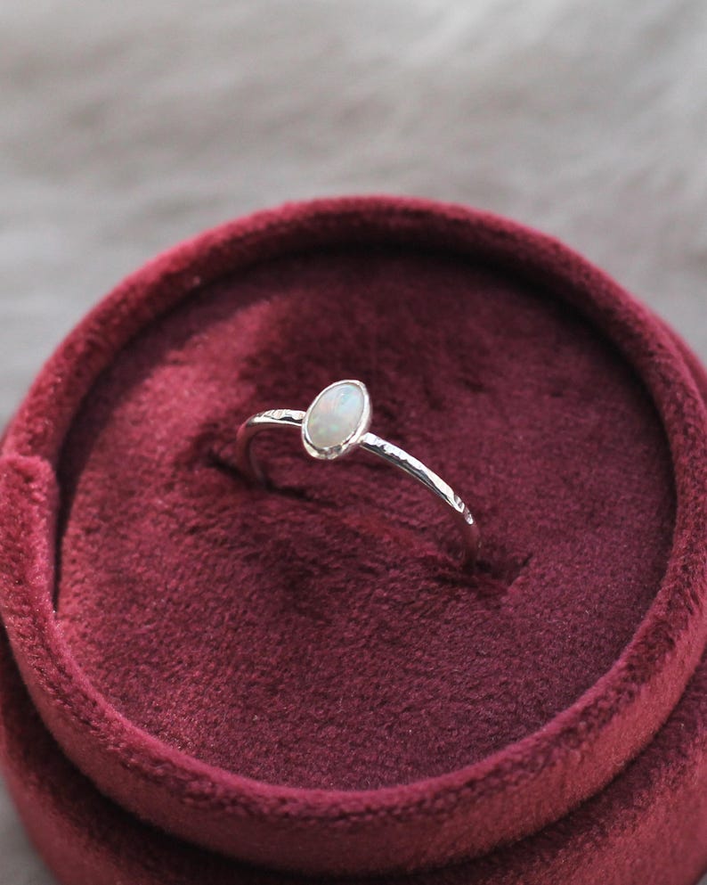 May include: A silver ring featuring a small, oval-shaped iridescent opal stone set in a smooth bezel. The ring band has a textured, hammered finish and is displayed upright in a deep red, circular velvet-lined jewelry box.