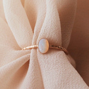 May include: A gold ring featuring a small, oval-shaped iridescent opal stone set in a bezel. The ring band has a distinct hammered, textured finish that catches the light, providing a reflective surface along the thin, metallic circular band.