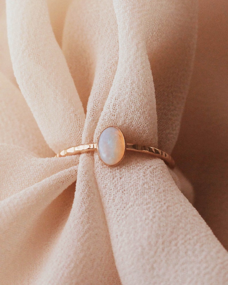 May include: A gold ring featuring a small, oval-shaped iridescent opal stone set in a bezel. The ring band has a distinct hammered, textured finish that catches the light, providing a reflective surface along the thin, metallic circular band.