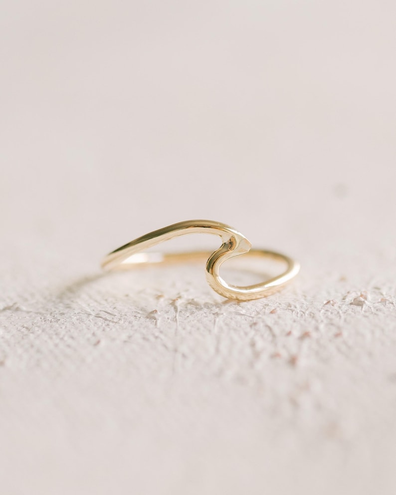 May include: A delicate gold ring with a wave design. The ring is made of polished gold and features a smooth, flowing shape. The ring is a minimalist piece of jewellery.