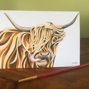 Puede incluir: Una pintura a la acuarela de una vaca de las Tierras Altas con pelaje largo y desgre&ntilde;ado de color marr&oacute;n claro y beige. La lengua de la vaca est&aacute; fuera. La obra de arte est&aacute; sobre un fondo blanco, con un pincel rojo sobre una superficie de madera.