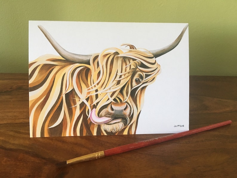 Puede incluir: Una pintura a la acuarela de una vaca de las Tierras Altas con pelaje largo y desgre&ntilde;ado de color marr&oacute;n claro y beige. La lengua de la vaca est&aacute; fuera. La obra de arte est&aacute; sobre un fondo blanco, con un pincel rojo sobre una superficie de madera.
