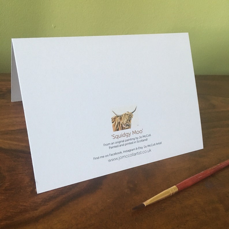 Puede incluir: Tarjeta de felicitaci&oacute;n blanca con el texto 'Squidgy Moo' y una ilustraci&oacute;n de una vaca de las Tierras Altas. La tarjeta tambi&eacute;n incluye el nombre del artista, la ubicaci&oacute;n y la direcci&oacute;n del sitio web. Un pincel descansa sobre una superficie de madera.