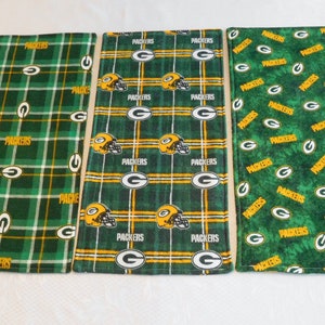 May include: Three green and gold fabric placemats with a repeating pattern of the Green Bay Packers logo. The placemats have different patterns, including a plaid pattern, a striped pattern, and a scattered pattern.