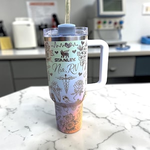 May include: A stainless steel Stanley tumbler with a white handle and a gradient color scheme. The tumbler is decorated with floral and medical-themed engravings, including the name "Nia, RN". The tumbler has a straw and a blue lid.