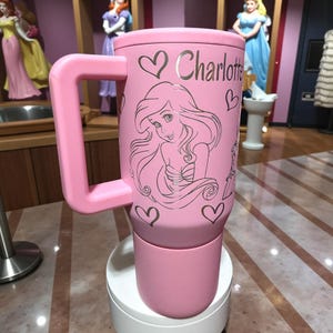 May include: A pink insulated travel mug with a large handle. The mug features a silver illustration of a mermaid, hearts, and the name "Charlotte". The mug is sitting on a white stand.