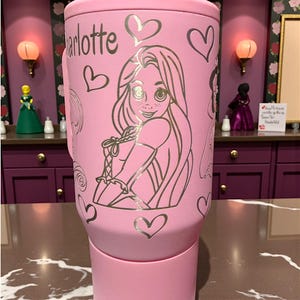 May include: Pink tumbler featuring a silver illustration of a princess, hearts, and the name "Charlotte." The tumbler has a handle and a lid. The design is a whimsical and decorative style.