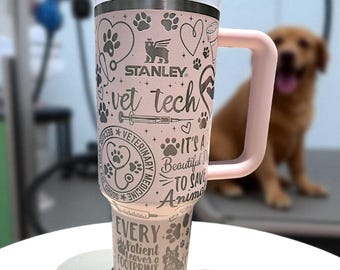 May include: A light pink Stanley tumbler with a handle and white lid, featuring black laser-etched designs including paw prints, hearts, a stethoscope, a syringe, and text reading vet tech, veterinary medicine, and every patient leaves a footprint.