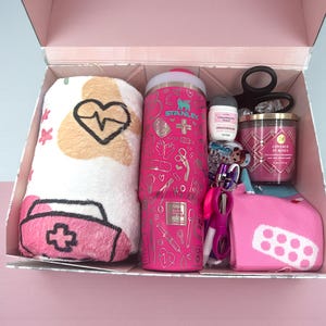 May include: A pink gift box filled with various items. Includes a plush blanket with a nurse's cap and heartbeat design, a pink Stanley tumbler, a small bottle of moisturizer, a candle, scissors, and a pink first-aid kit.