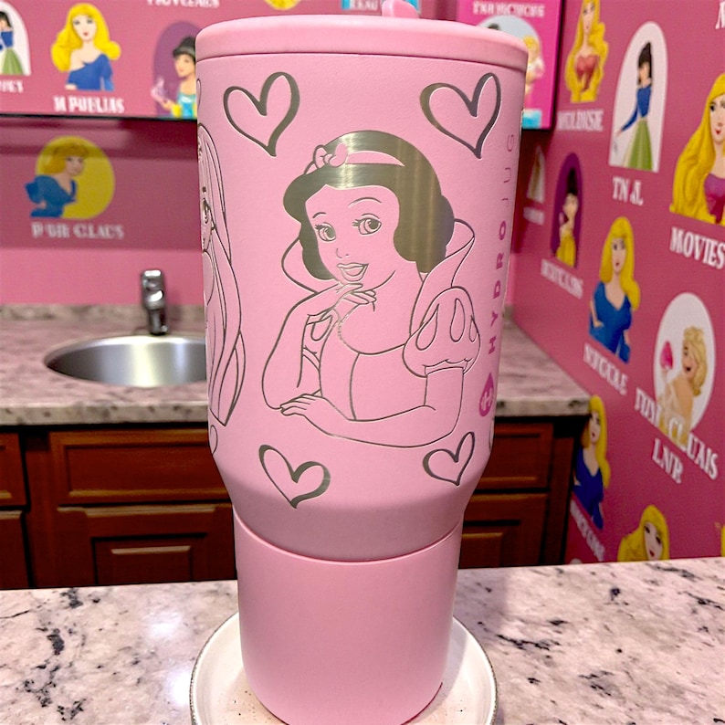 May include: A pink insulated tumbler featuring a Snow White design with hearts. The tumbler has a silver outline of Snow White and hearts. The tumbler is sitting on a white plate.