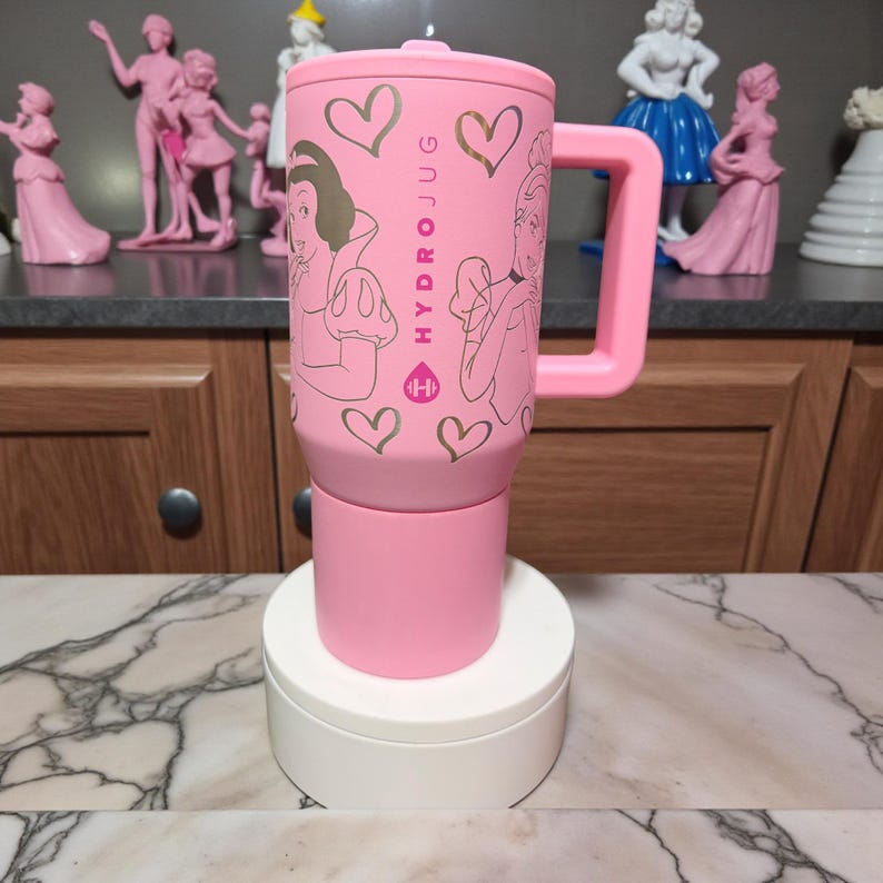 May include: A pink HydroJug water bottle with a handle, featuring illustrations of princesses and gold heart accents. The bottle has the text "HYDROJUG" printed on it.