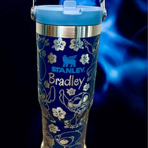 May include: A blue stainless steel travel mug with a blue lid and handle. The mug has a gold design of Stitch from Lilo & Stitch, flowers, and the Stanley logo. The name "Bradley" is engraved on the mug.