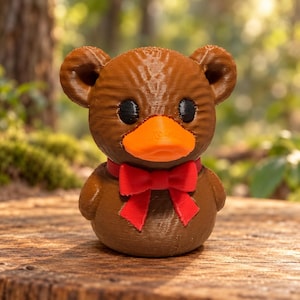 May include: A brown bear-shaped figurine with a bright orange duck-like bill, small black eyes, and a prominent red bow tie around its neck. The figure has a textured surface and rounded ears, standing in a compact, upright posture.