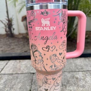 May include: A pink Stanley tumbler with a handle and lid. The tumbler features a gradient design and is adorned with illustrations of characters and the name "Angela". The Stanley logo is visible.