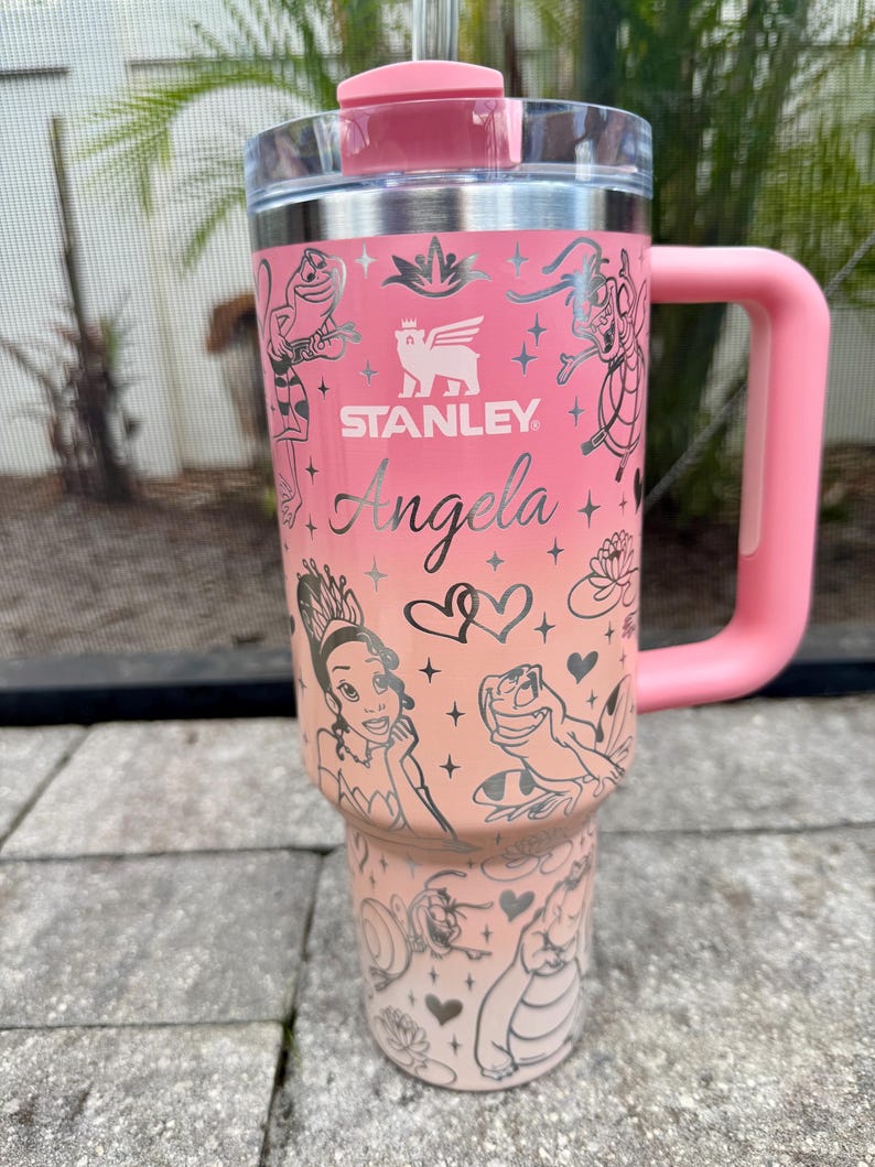 May include: A pink Stanley tumbler with a handle and lid. The tumbler features a gradient design and is adorned with illustrations of characters and the name "Angela". The Stanley logo is visible.