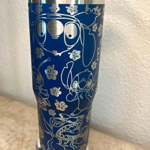 May include: A blue stainless steel tumbler with a silver lid and a silver base. The tumbler features a design of Stitch and other characters from the Disney movie Lilo & Stitch. The design is engraved in silver.