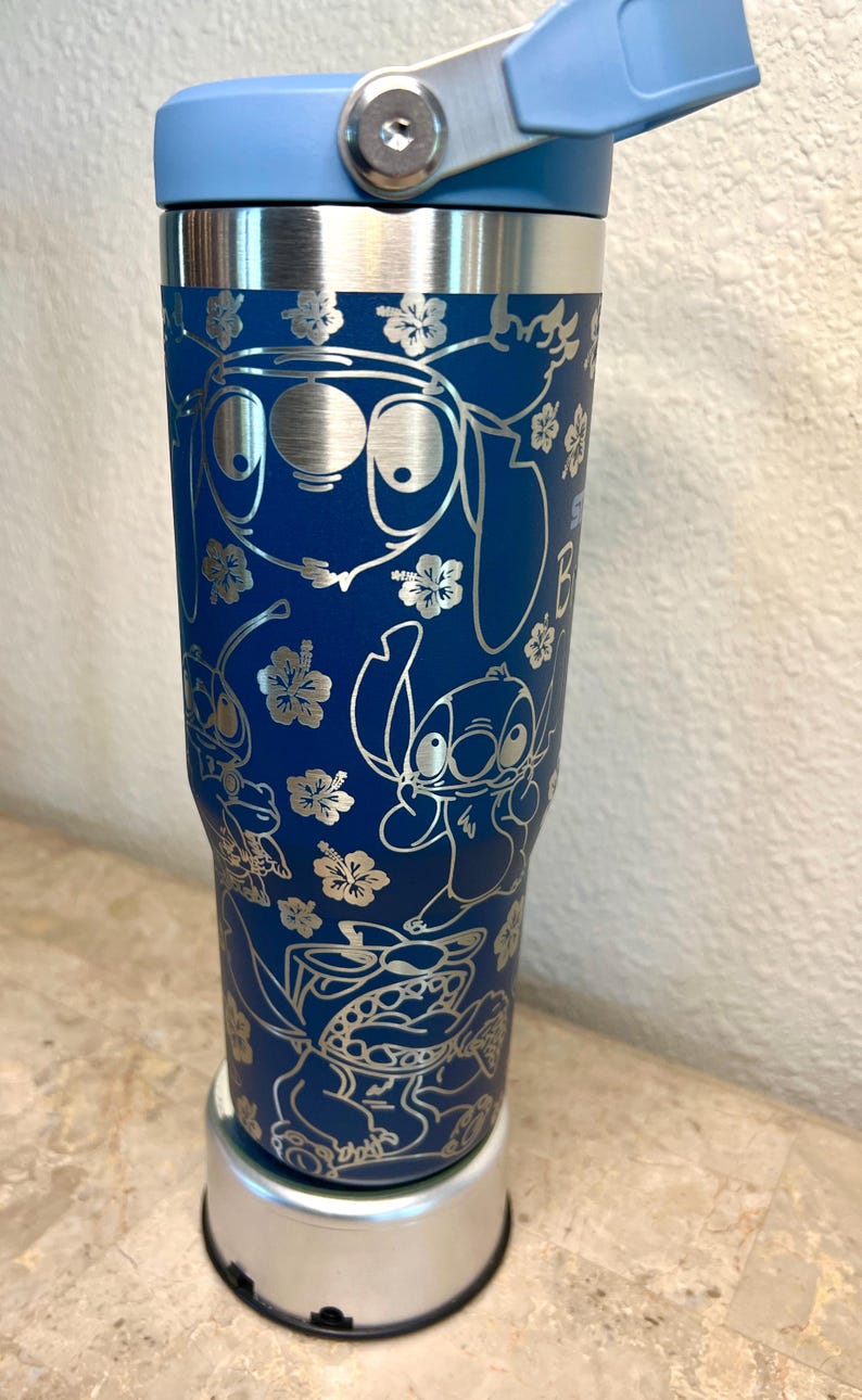 May include: A blue stainless steel tumbler with a silver lid and a silver base. The tumbler features a design of Stitch and other characters from the Disney movie Lilo & Stitch. The design is engraved in silver.