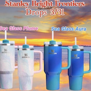 May include: Four Stanley tumblers with handles and straws are displayed. Two are blue, one is white with a pink handle, and one is white with a blue handle. The text "Stanley Bright Frontiers Drops 3/31" is at the top.