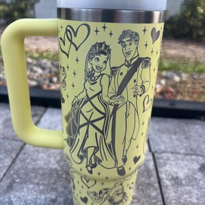 May include: A yellow travel mug with a handle, featuring a black illustration of a princess and prince. The mug has a white lid, a yellow straw, and is decorated with hearts and other characters. The mug is designed for cold beverages.