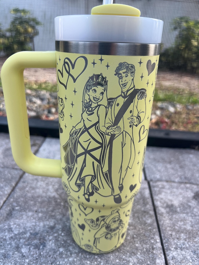 May include: A yellow travel mug with a handle, featuring a black illustration of a princess and prince. The mug has a white lid, a yellow straw, and is decorated with hearts and other characters. The mug is designed for cold beverages.