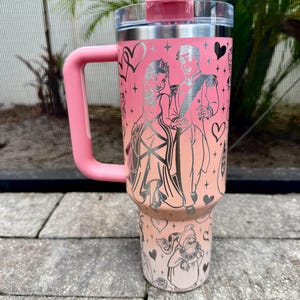 May include: A pink insulated tumbler with a handle, lid, and straw. The tumbler features a design of a couple in formal attire, hearts, and other illustrations. The handle and lid are a matching pink color.