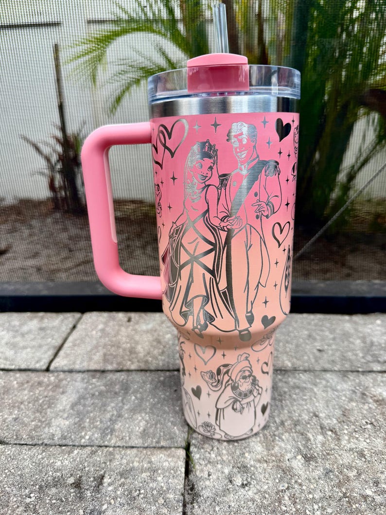 May include: A pink insulated tumbler with a handle, lid, and straw. The tumbler features a design of a couple in formal attire, hearts, and other illustrations. The handle and lid are a matching pink color.