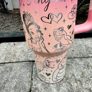 May include: A pink tumbler with a handle, featuring the name "Angela" in silver script. The tumbler is decorated with illustrations of a princess, a frog, hearts, and stars. The design is a gradient from light pink to a darker pink.