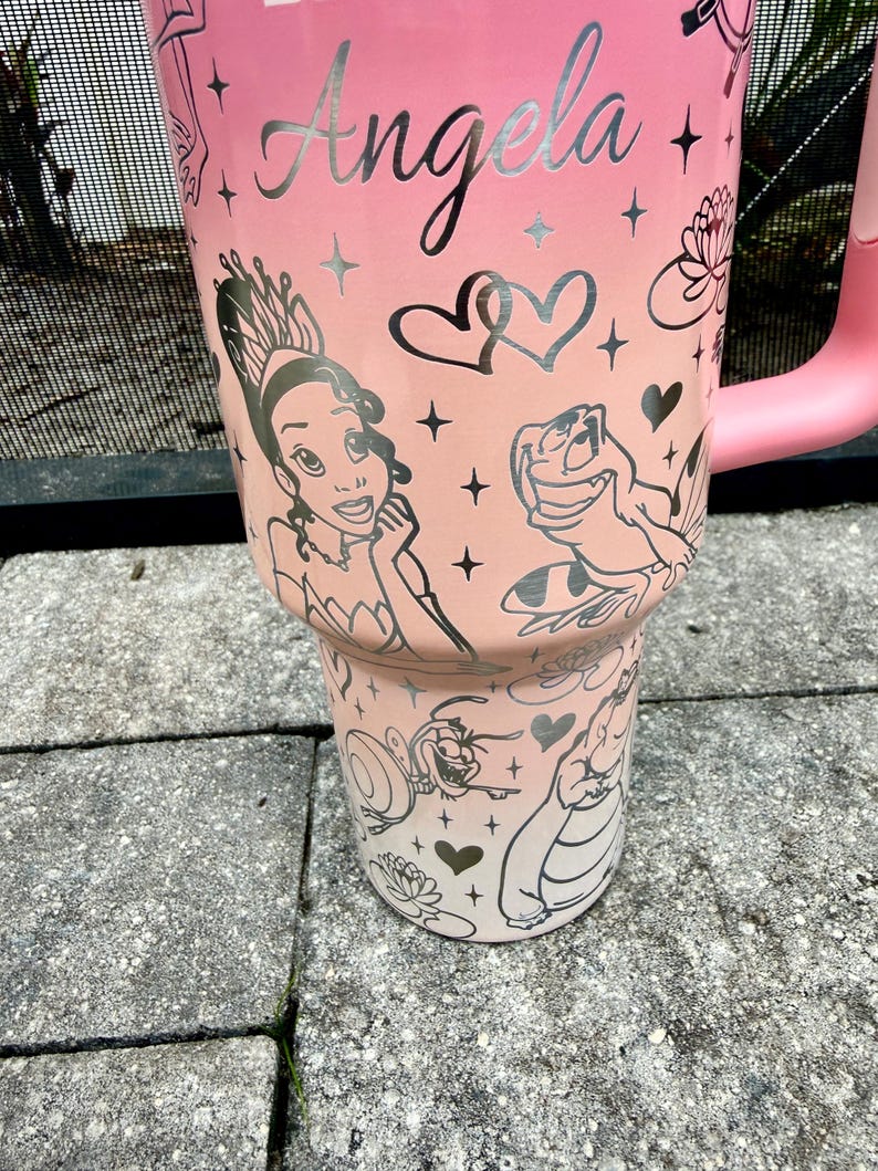 May include: A pink tumbler with a handle, featuring the name "Angela" in silver script. The tumbler is decorated with illustrations of a princess, a frog, hearts, and stars. The design is a gradient from light pink to a darker pink.