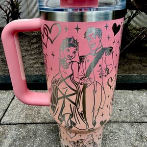 May include: Pink travel tumbler with a handle and a lid. The tumbler features a princess and prince design with hearts and stars. The handle and lid are a matching pink color. The tumbler is made of stainless steel.