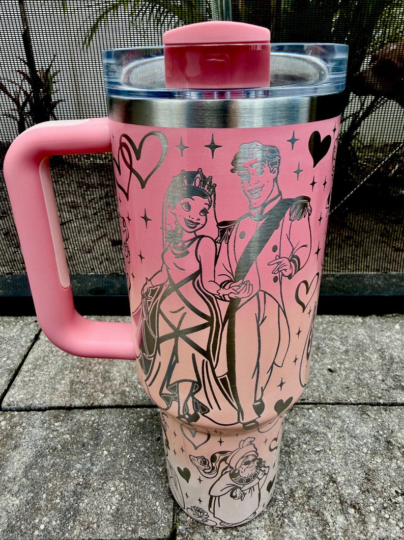 May include: Pink travel tumbler with a handle and a lid. The tumbler features a princess and prince design with hearts and stars. The handle and lid are a matching pink color. The tumbler is made of stainless steel.