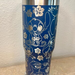 May include: A blue stainless steel travel mug with a blue lid and a silver base. The mug features a white design of Stitch from Lilo & Stitch, with a tropical floral pattern.