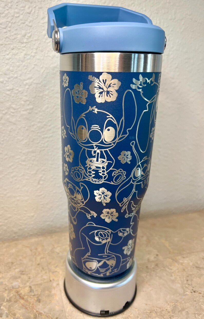May include: A blue stainless steel travel mug with a blue lid and a silver base. The mug features a white design of Stitch from Lilo & Stitch, with a tropical floral pattern.