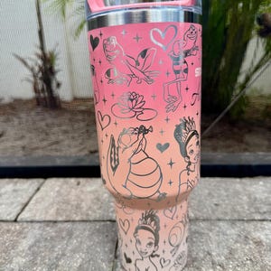 May include: A stainless steel tumbler with a pink and peach gradient design. The tumbler features silver illustrations of frogs, a water lily, and a princess. The tumbler has a clear lid and a pink handle.
