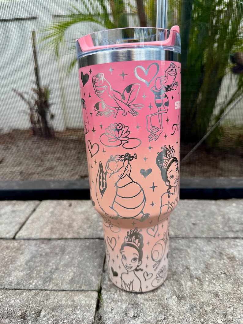 May include: A stainless steel tumbler with a pink and peach gradient design. The tumbler features silver illustrations of frogs, a water lily, and a princess. The tumbler has a clear lid and a pink handle.