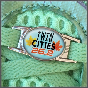 Könnte beinhalten: Ein silberner Schnallenschmuck mit blauem Hintergrund und dem Text "Twin Cities 26.2" in Schwarz und Orange. Der Schmuck zeigt drei orangefarbene und gelbe Ahornblätter.