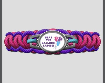 May include: A braided paracord bracelet featuring a central oval metal charm. The cord is woven in alternating bright pink and deep purple strands. The charm displays the text BEAT THE BALLOON LADIES! with a graphic of three mouse-shaped balloons.