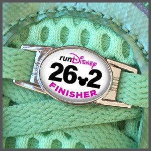 Può includere: Un ciondolo per scarpe argentato con sfondo bianco e testo nero che recita "runDisney 26.2 FINISHER" con una testa di Topolino al centro del numero 2.