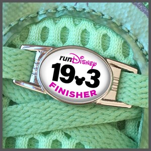Può includere: Un ciondolo per scarpe argentato con un'etichetta ovale bianca che dice "runDisney 19*3 FINISHER" in lettere nere e rosa. Il ciondolo è attaccato a uno stringa di scarpa verde.