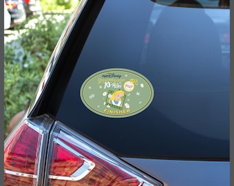 May include: An oval-shaped sticker on a car window featuring a light green background with the text runDisney Springtime Surprise 10-Miler 2027 Finisher. The design includes an illustration of Alice, a pocket watch, and small white flower accents.