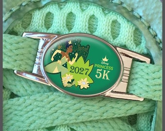 May include: An oval metal shoe lace charm featuring a green background with an illustration of Tiana in a green dress, yellow flowers, and the text Disney Princess 2027 5K. The charm is attached to light green woven shoe laces with a mesh texture.