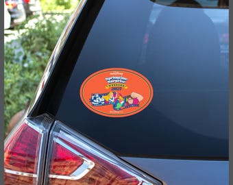 May include: An oval-shaped orange sticker applied to a car window featuring the text Springtime Surprise Weekend 2027. The design includes a film strip graphic with illustrations of Mickey Mouse, Alice, Peter Pan, and Wreck-It Ralph characters.