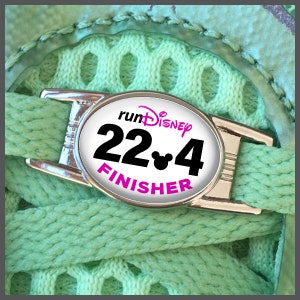 Può includere: Un ciondolo per scarpe ovale bianco con testo nero che recita "runDisney 22.4 FINISHER" e un'icona di testa di Topolino nera.