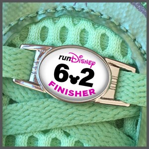 Può includere: Un ciondolo per scarpe argentato con un'etichetta ovale bianca che dice "runDisney 6.2 FINISHER" in testo nero con una testa di Topolino rosa tra i numeri.