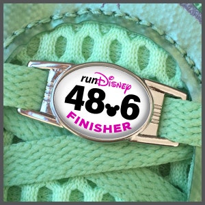 May include: A white oval-shaped running shoe charm with a black and white Mickey Mouse head logo and the text "runDisney 48.6 FINISHER" in black text with a pink outline.