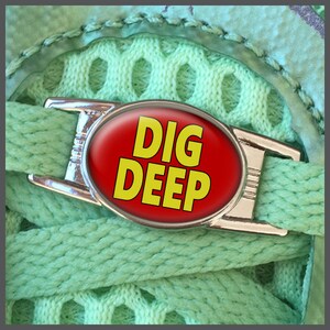 Op de afbeelding: Een zilveren veterscharm met een rode ovale achtergrond en de woorden "DIG DEEP" in gele letters.
