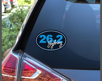 May include: A blue and black oval sticker with the text "26.2 Sydney" on a car window.